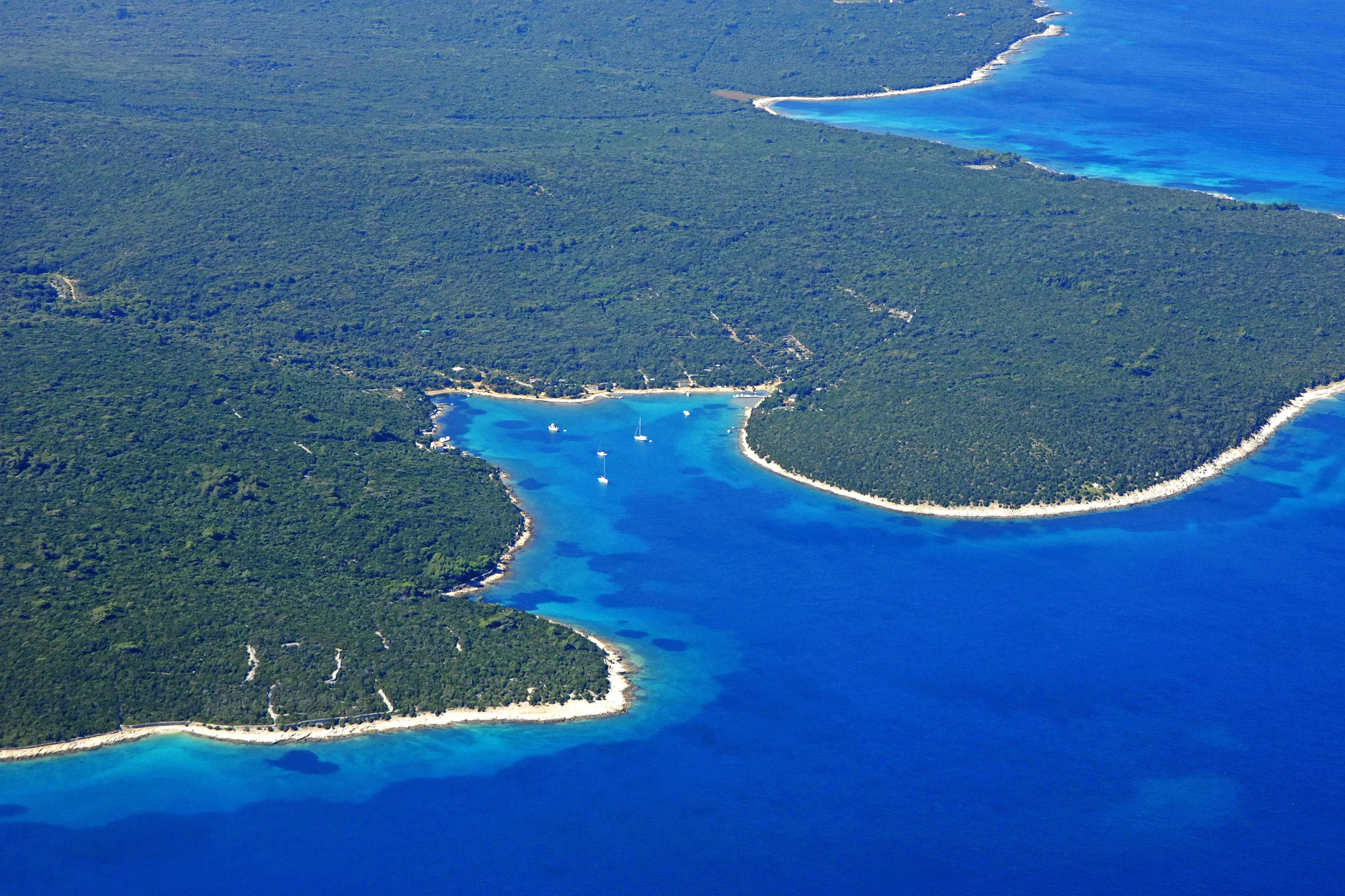 Aerial of forested Adriatic island coastline with sheltered bay and few anchored sailboats