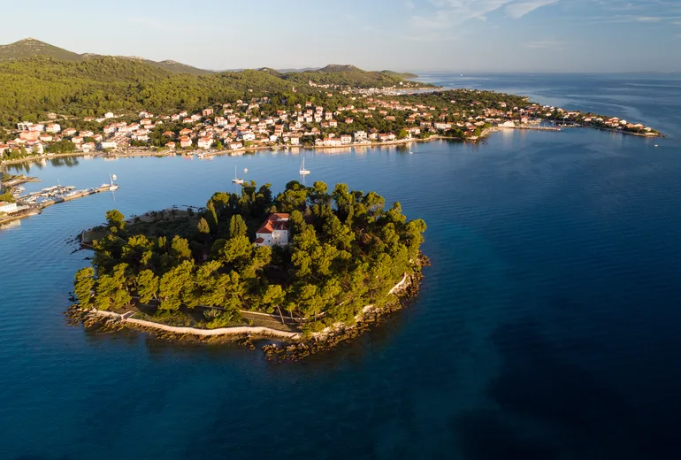 Golden-hour aerial of a small wooded islet with a monastery and a coastal village behind
