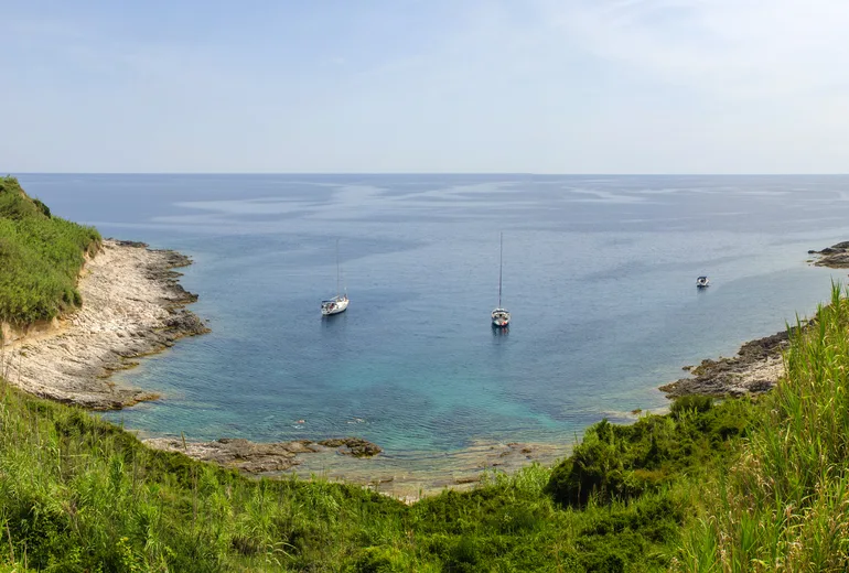 Three sailboats anchored in a quiet green-framed rocky cove on a hazy summer day