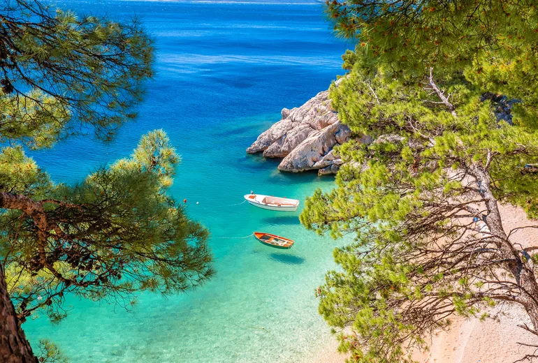 Turquoise cove with a pebble beach and two small boats framed by pine trees